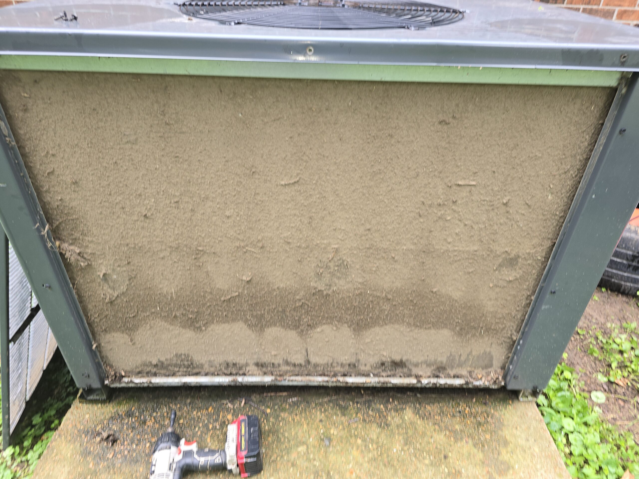 Dirty air conditioning unit with dusty coils, placed on a concrete slab outdoors. A power drill lies on the ground in front.