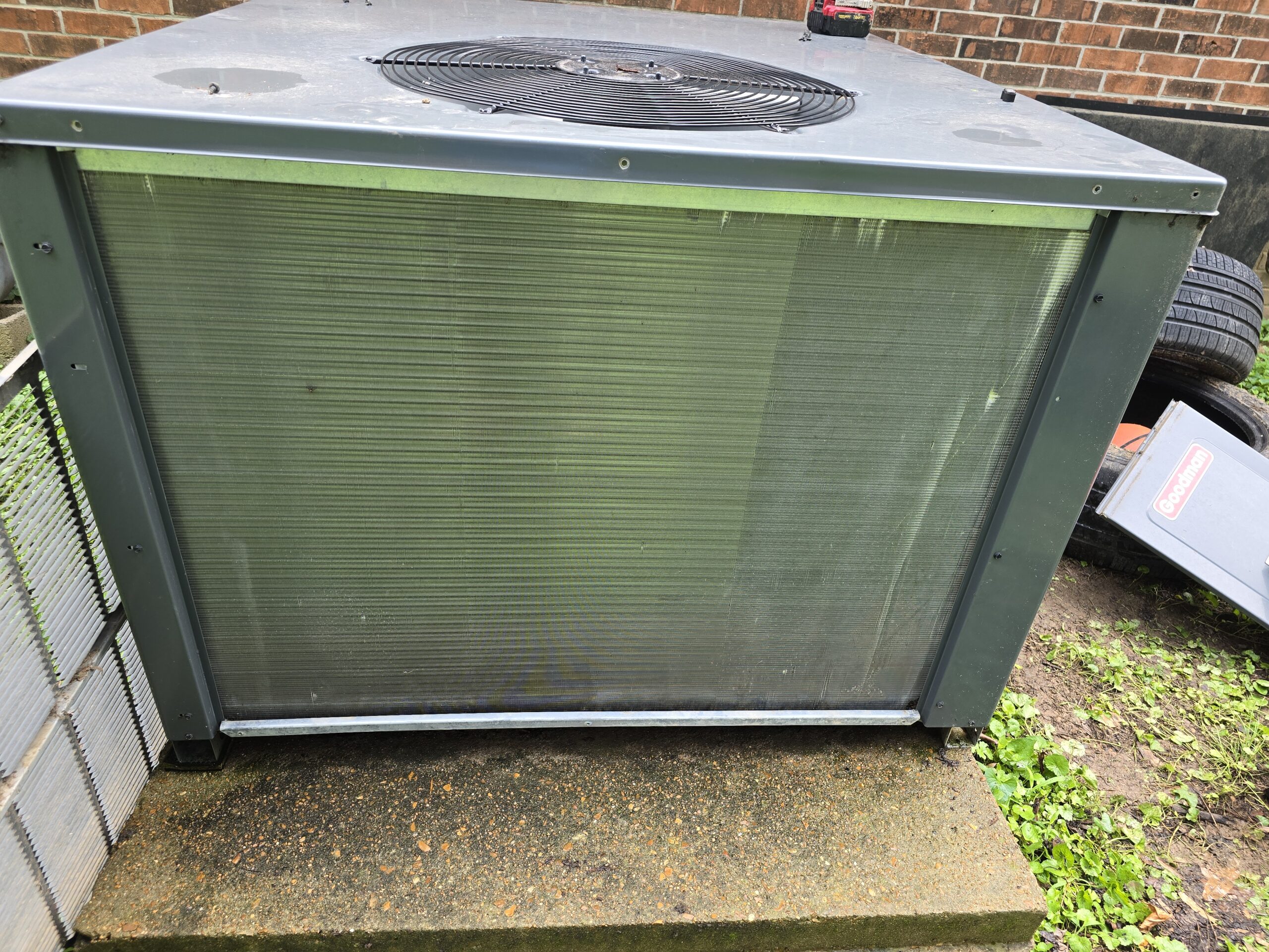 An outdoor HVAC unit on a concrete pad next to a brick wall and grass. A tire and tool are nearby.