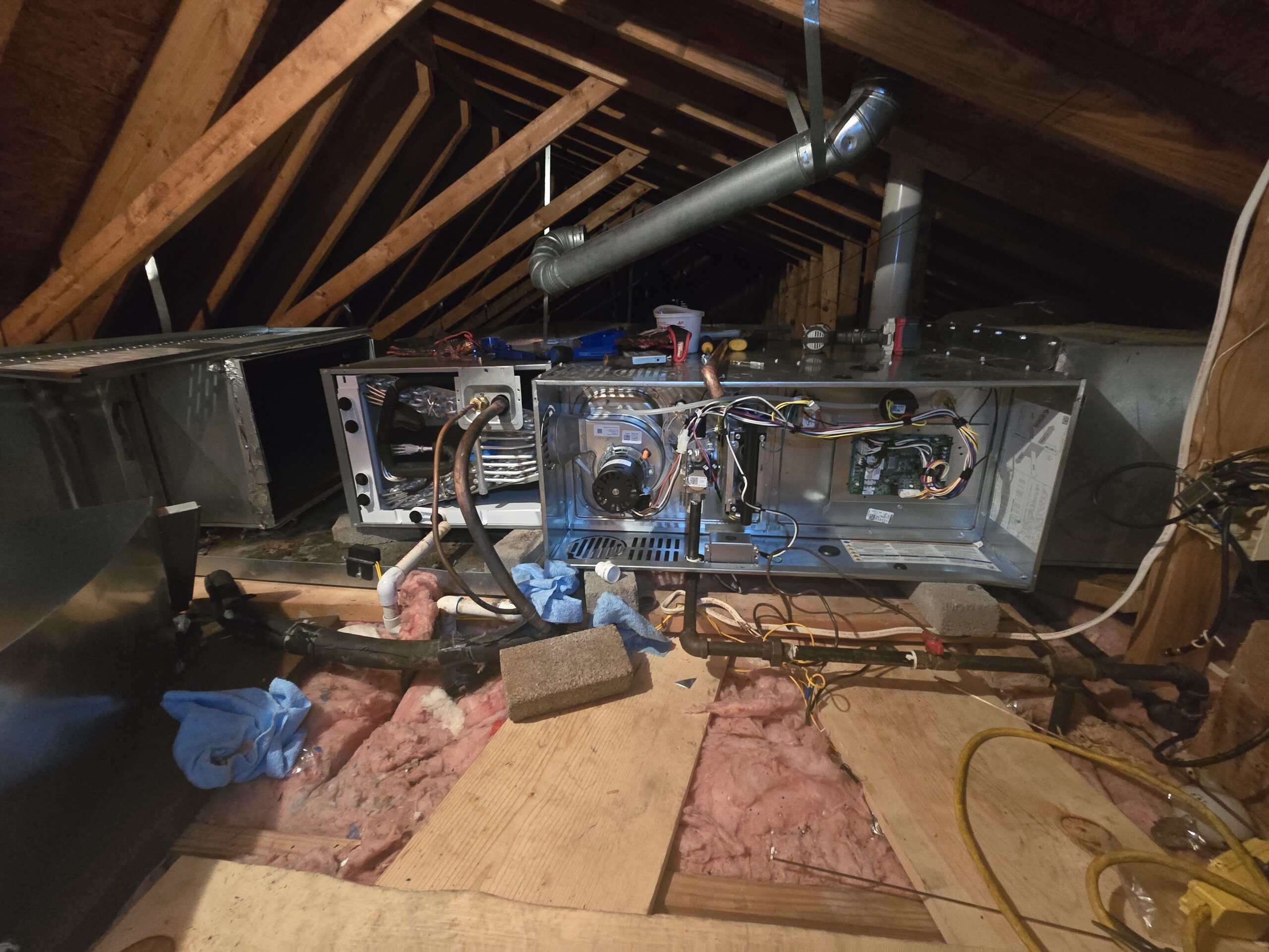 An open HVAC system is exposed in an attic, surrounded by insulation and wooden beams, with various tools and materials scattered around.