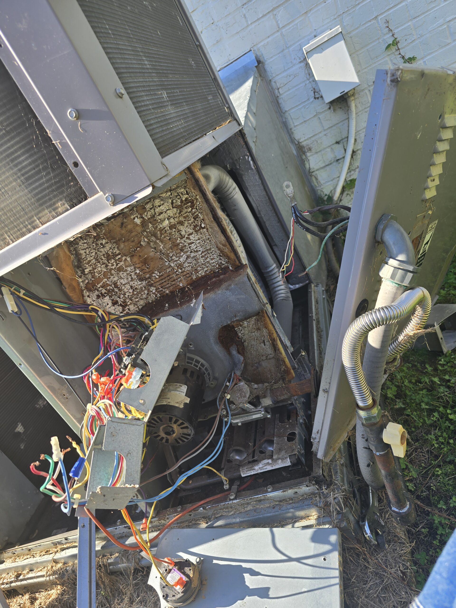 Open HVAC unit with exposed wires and components, situated outside against a brick wall.