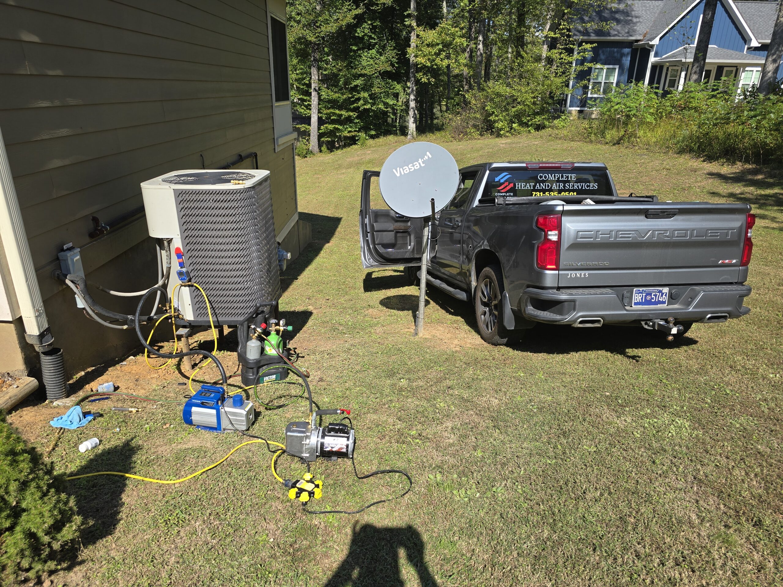 A truck parked next to a house with HVAC equipment and tools set up on the grass for maintenance work. A satellite dish is visible nearby.