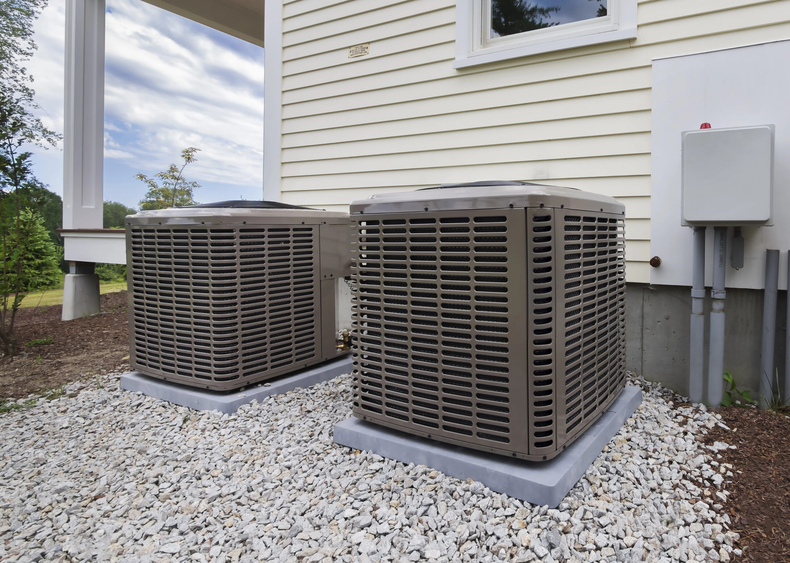 Two outdoor air conditioning units, expertly installed, rest on a gravel bed next to a building with beige siding.