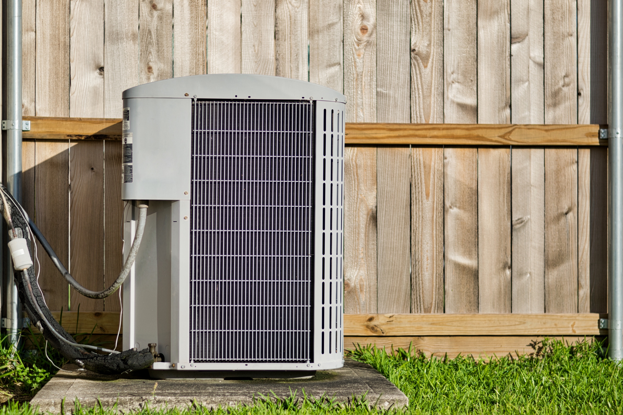 An AC installation showcases an outdoor air conditioning unit perched on a concrete slab in front of a wooden fence, nestled amidst lush grass.