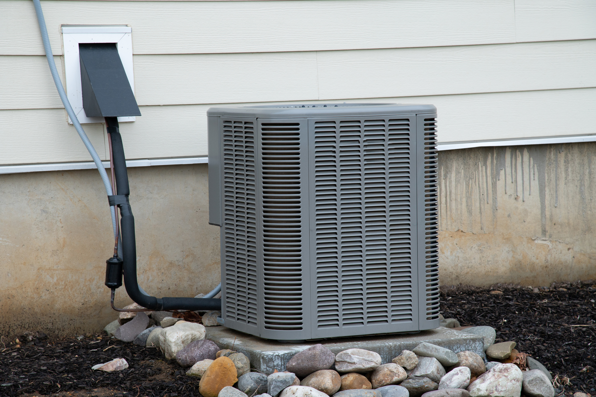 A gray Heat pump unit, part of a comprehensive heating installation, is mounted outside against a beige wall, nestled among small rocks and mulch.